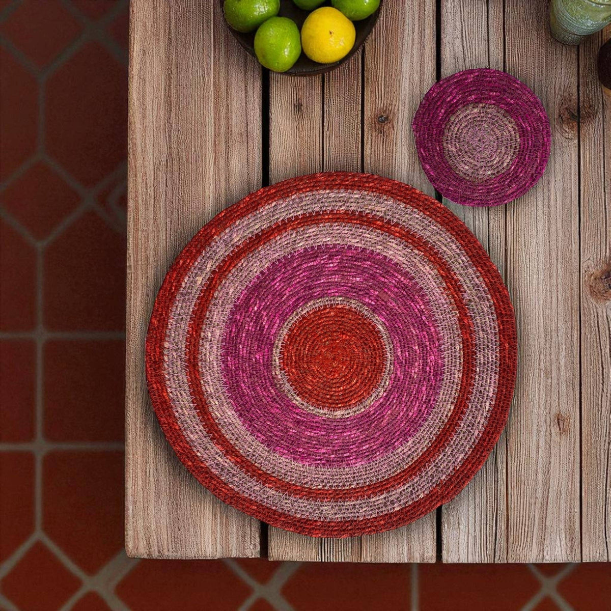 A round woven placemat with concentric rings of red, pink, and beige sits on a rustic wooden table, with a matching small round coaster above it and limes in the corner.
