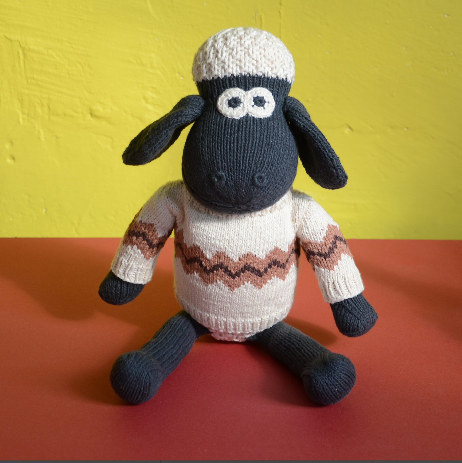 A knitted sheep doll with a black body and face, wearing a cream-coloured sweater with a brown zigzag pattern and a cream knit hat, sitting against a yellow and orange background.