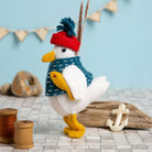 A white felt seagull toy wearing a red and blue hat, holding a yellow felt fish under their wing, surrounded by nautical items and colours.