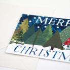 A close-up of a Christmas greeting card featuring a textured, collage-style winter forest with green and yellow trees under a dark blue sky, with the words 'MERRY CHRISTMAS' partially visible.