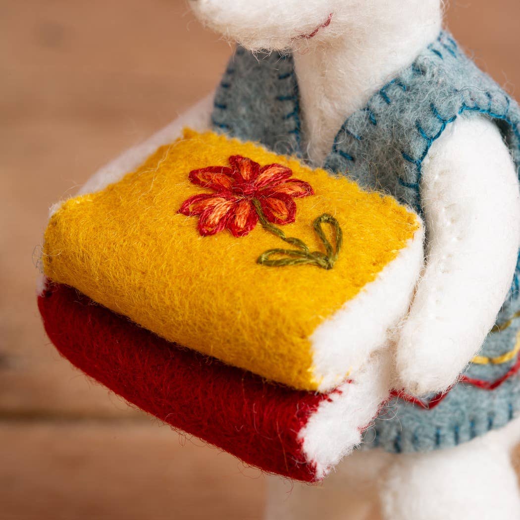 White plush toy holding a yellow book with a red flower design on a blurred background