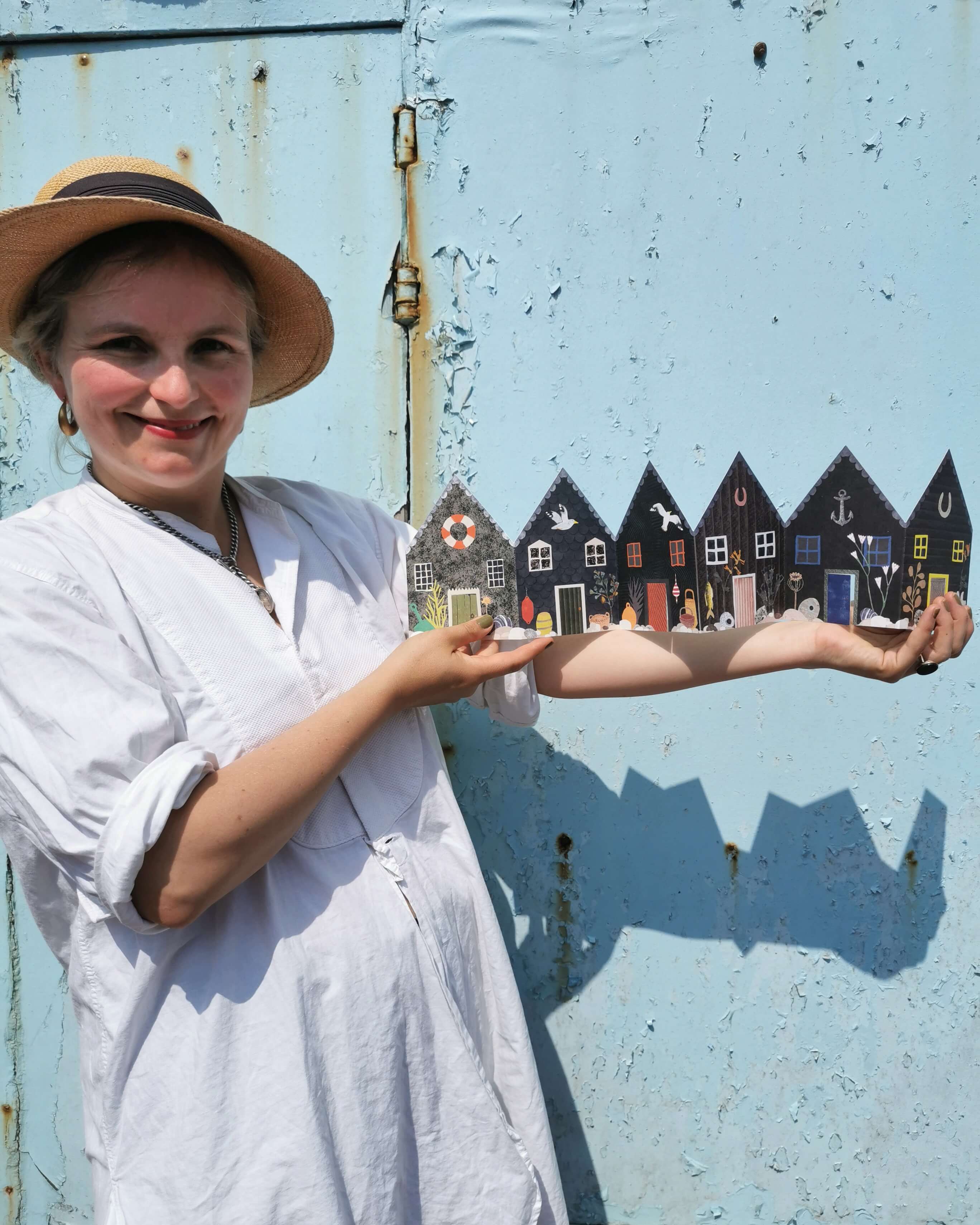 Woman in a white dress and hat standing in front of a blue wall with a small village mural.