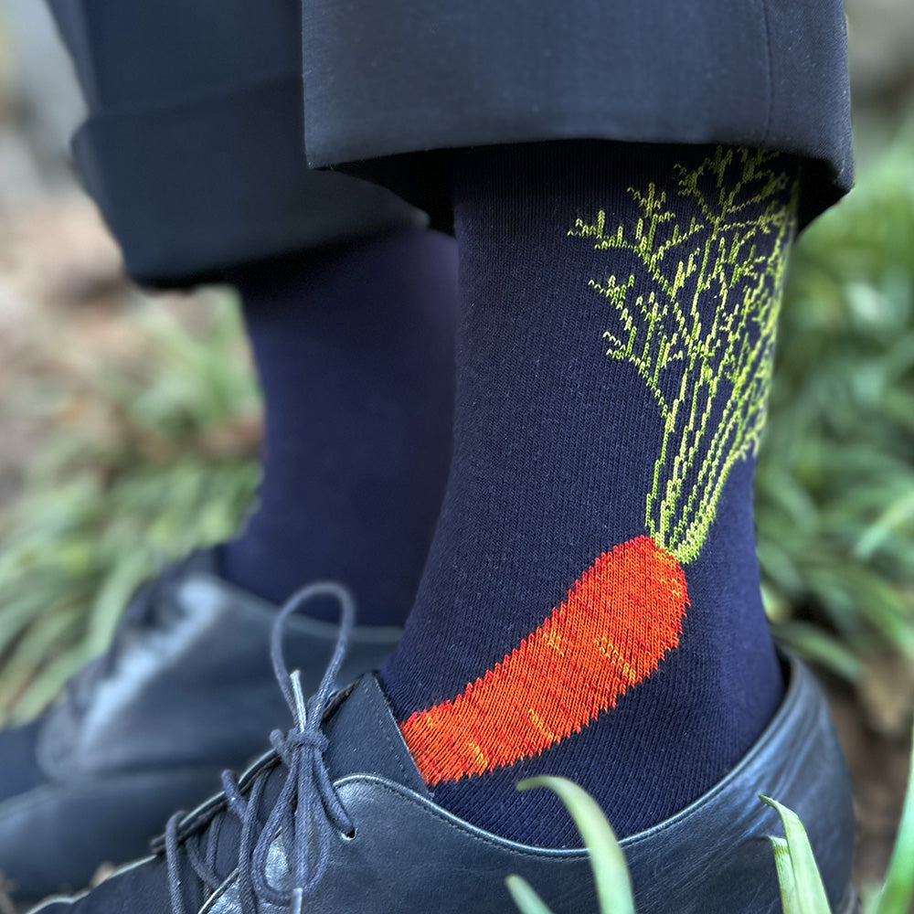 A close-up of a person's legs wearing navy blue socks with a large orange carrot and green leafy tops printed on one sock, paired with black dress shoes and dark trousers in an outdoor setting.