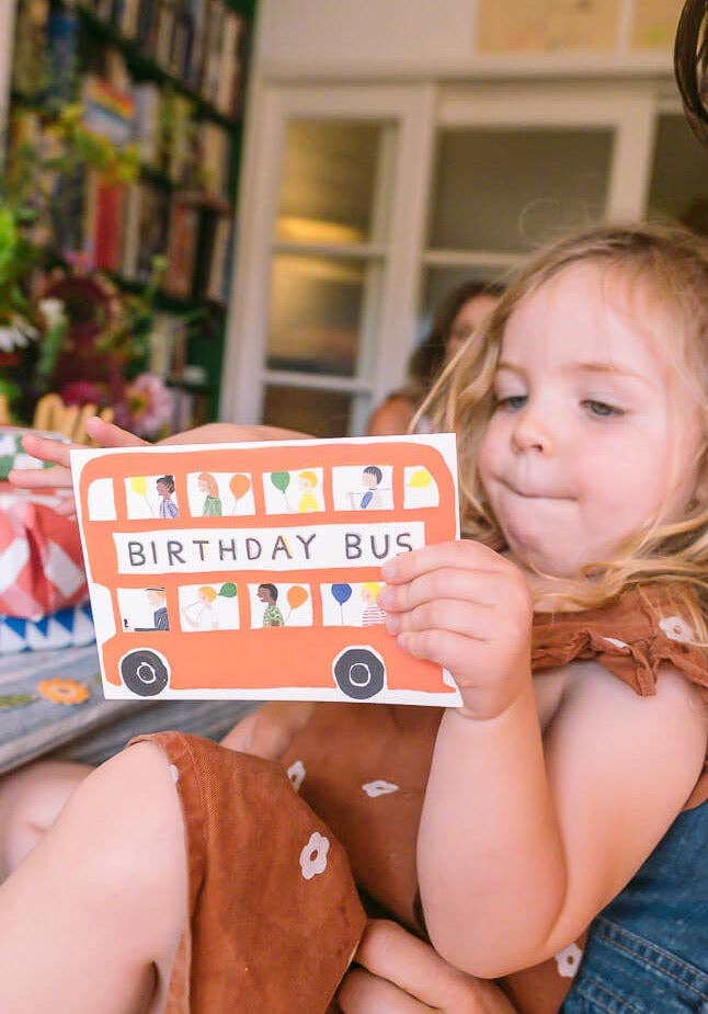 Child holding a card with 'Birthday Bus' text in a cozy indoor setting.