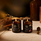 Two lit candles in amber jars on a wooden surface with decorative elements.