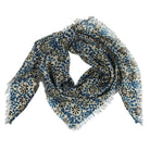 A square scarf with fringed edges, featuring a blue and black floral block-print pattern on a cream background, arranged in a loose knot on a light gray studio background.