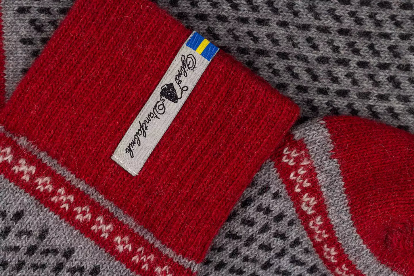 Close-up of a red knit sock with a white brand tag  and a small Swedish flag, laid over a gray and black patterned knit textile.