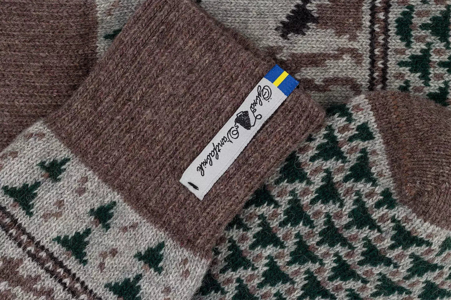 Close-up of a knitted sock with a brown ribbed cuff featuring a white tag that reads 'Guld Handgross' with a small blue and yellow accent, over a patterned scarf in brown, beige, and green.